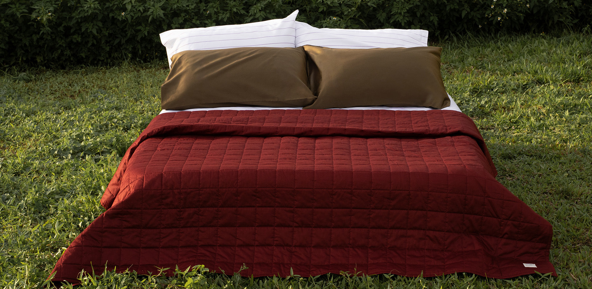 Red lightweight quilt blanket with moss green pillows on a bed outdoors in a grassy area
