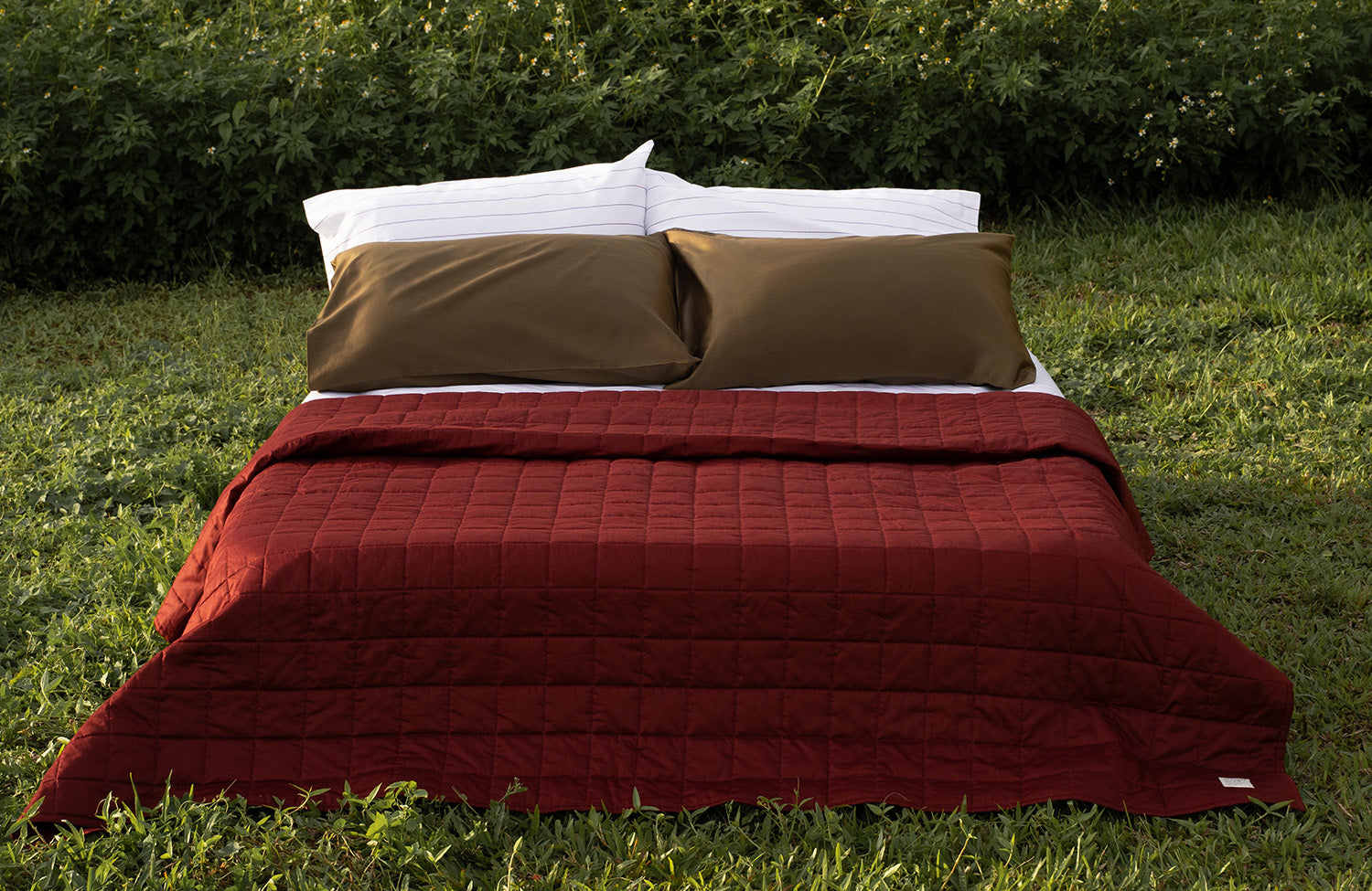 Red lightweight quilt blanket and moss green pillows on a bed outdoors with green grass and bushes in the background